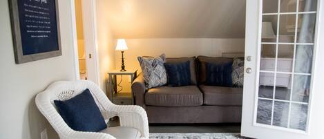 A cozy living room with a white wicker chair, a brown sofa with patterned cushions, a small side table with a lamp, and a patterned rug.