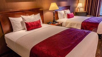 A hotel room with two beds, each with white linens and red decorative pillows. There are bedside tables with lamps and a telephone.