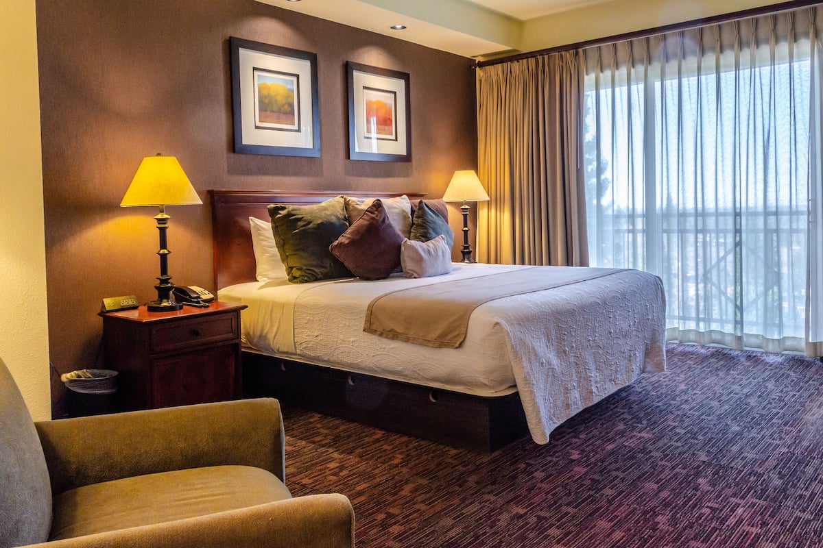 Habitación de hotel con una cama grande, dos lámparas de noche, una mesita de madera, una silla y un ventanal con cortinas.