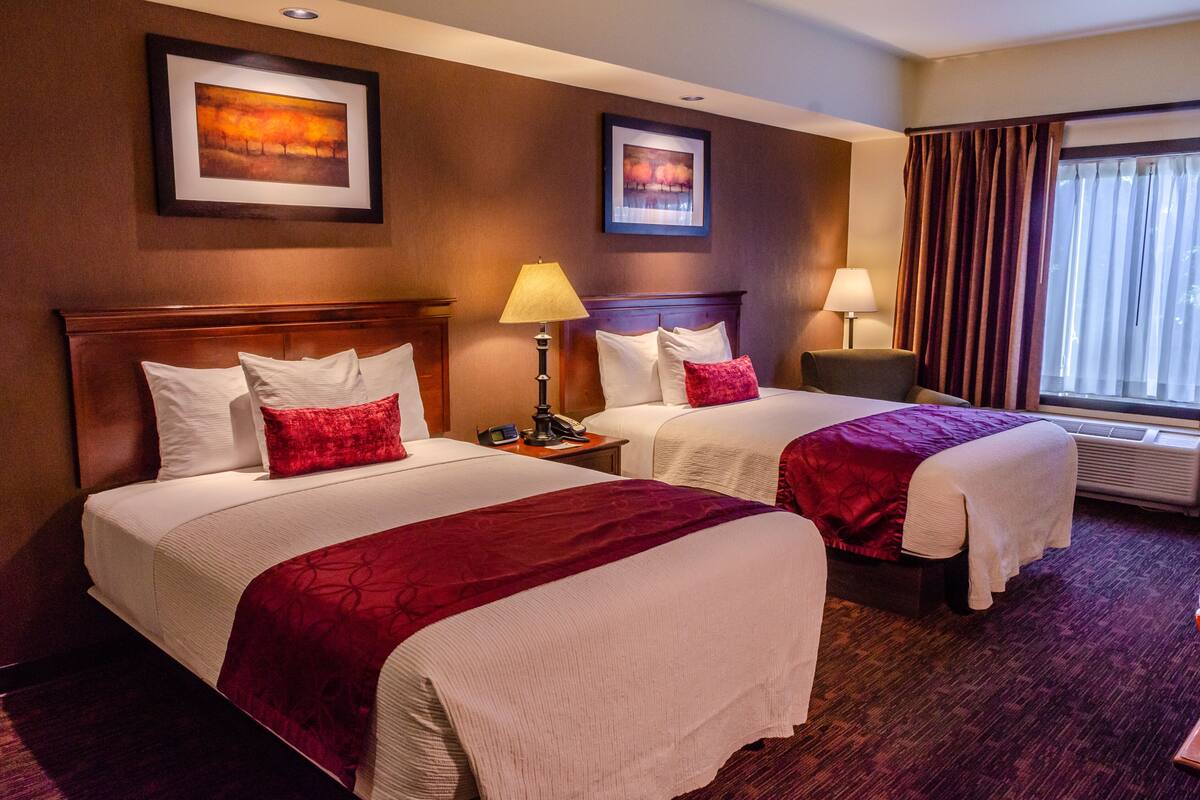 Habitación de hotel con dos camas, cada una con colcha roja y blanca, una mesita de noche con lámpara y un cuadro en la pared.
