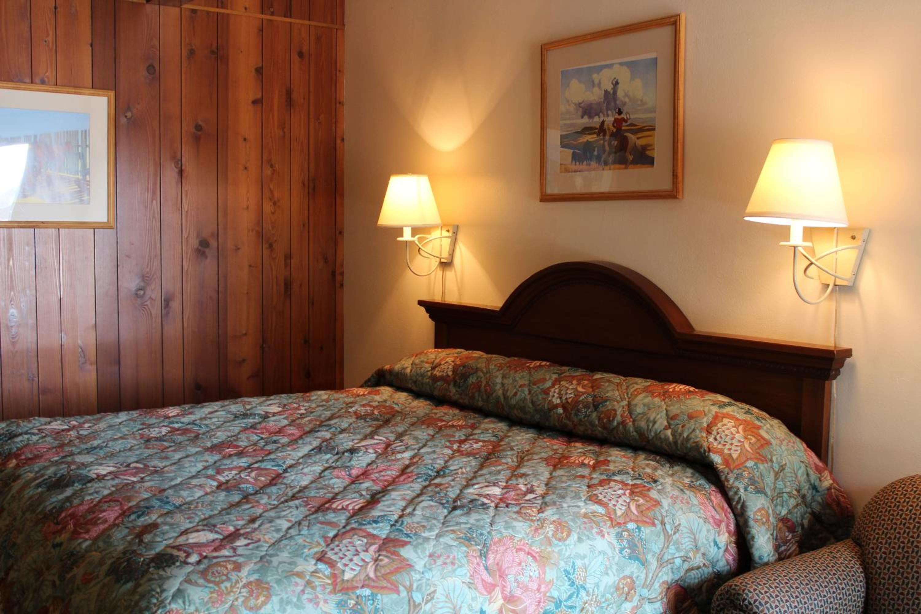 A hotel room with a wooden headboard, two wall-mounted lamps, a bed with a floral bedspread, and a framed picture on the wall.