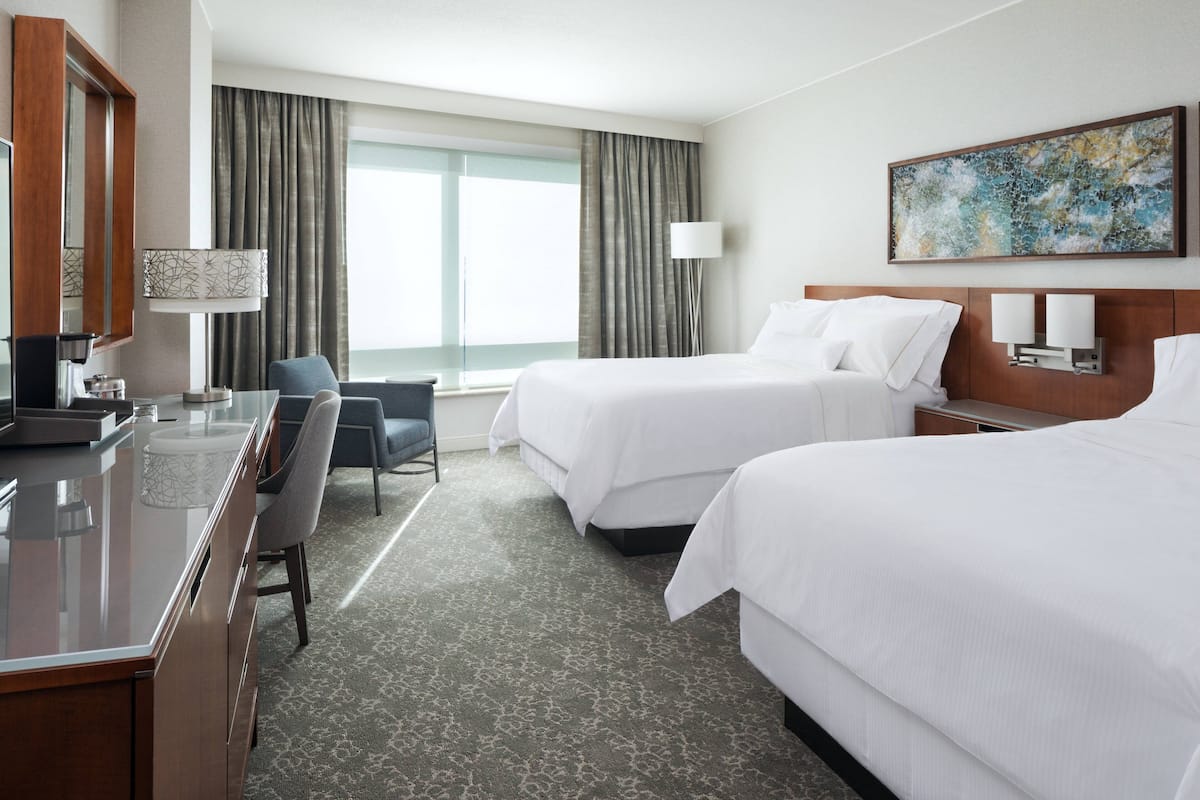 A hotel room with two beds, a desk, a chair, a lamp, and a large window with curtains.