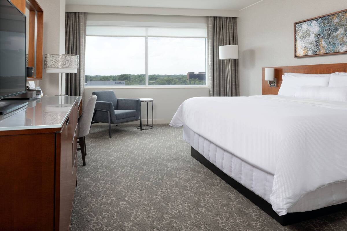 A hotel room with a large bed, a desk with a television, a chair, a small table, and a window with curtains.
