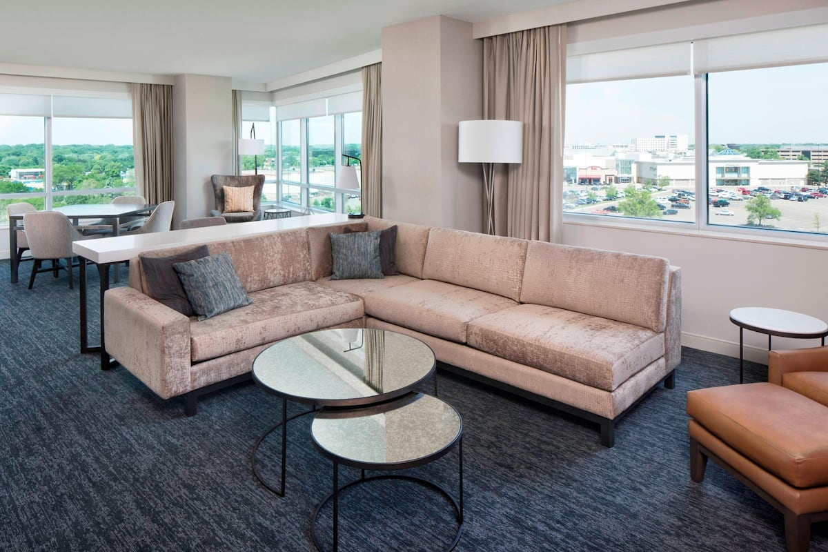 A hotel room with a large window, a sectional sofa, a dining table, and two small side tables.