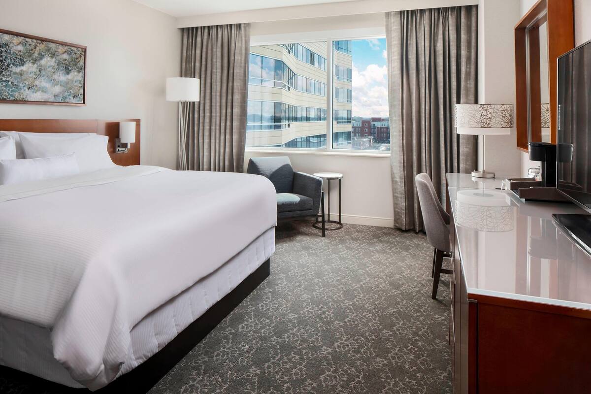 A hotel room with a large bed, a desk with a chair, a small table, a TV, and a window with curtains.