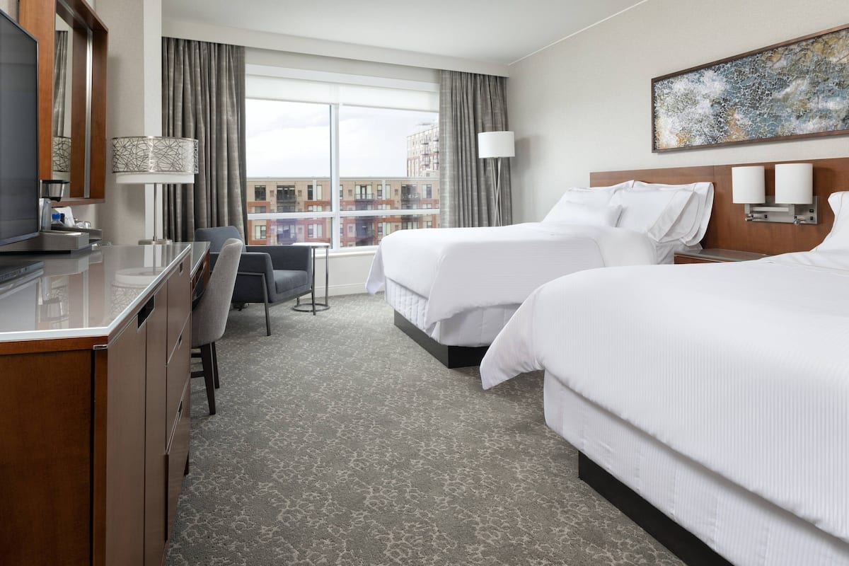 A hotel room with two beds, a desk, a chair, a TV, and a large window with curtains.