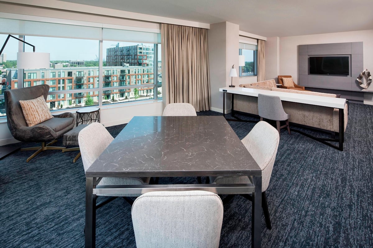 A modern hotel room with a large window offering a city view, a dining area with a table and chairs, and a flat-screen TV mounted on the wall.