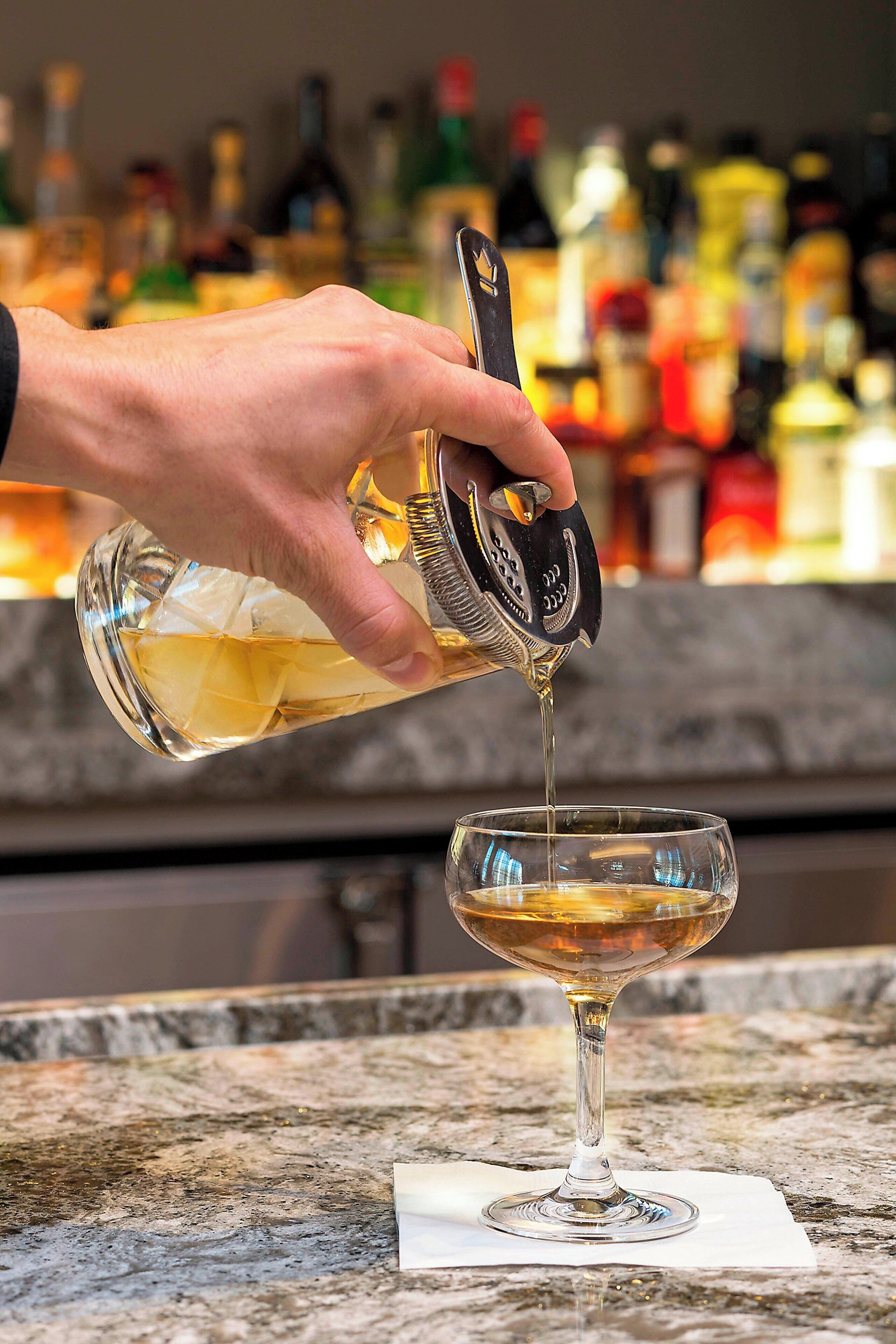 Eine Hand gießt eine goldfarbene Flüssigkeit aus einer Flasche in ein Glas auf einer Marmorplatte.