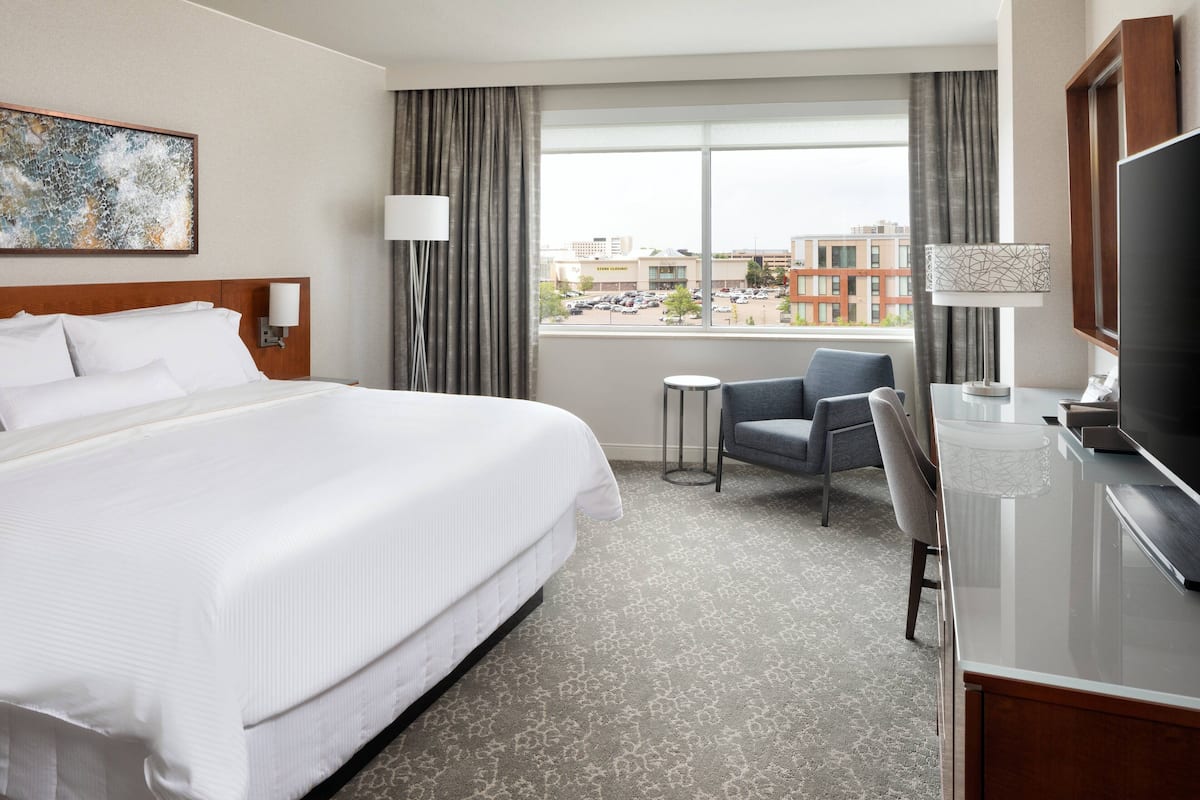 A hotel room with a large bed, a desk with a TV, a chair, a small table, and a window with a city view.