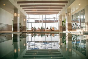 Amplia zona de piscina interior con reflejos nítidos, ventanas grandes y un área de descanso con plantas.