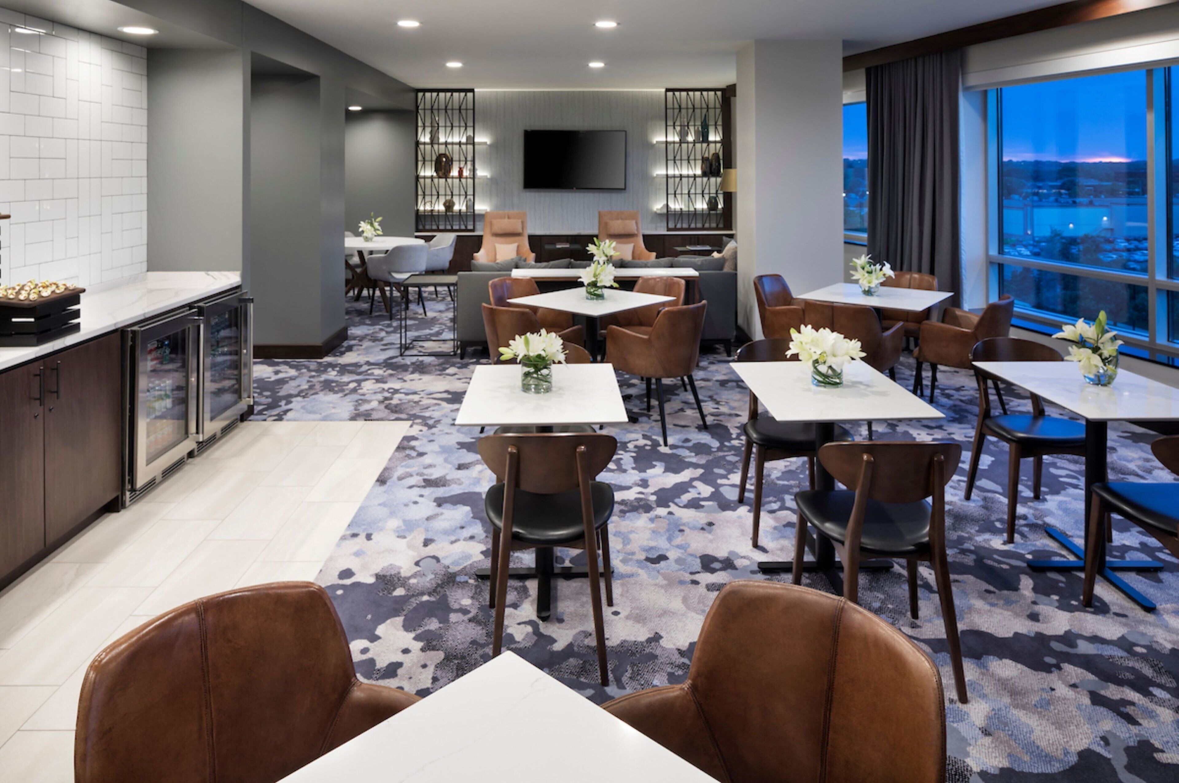 Un comedor moderno de hotel con mesas y sillas, una zona de cocina y un amplio ventanal con vistas a la ciudad.