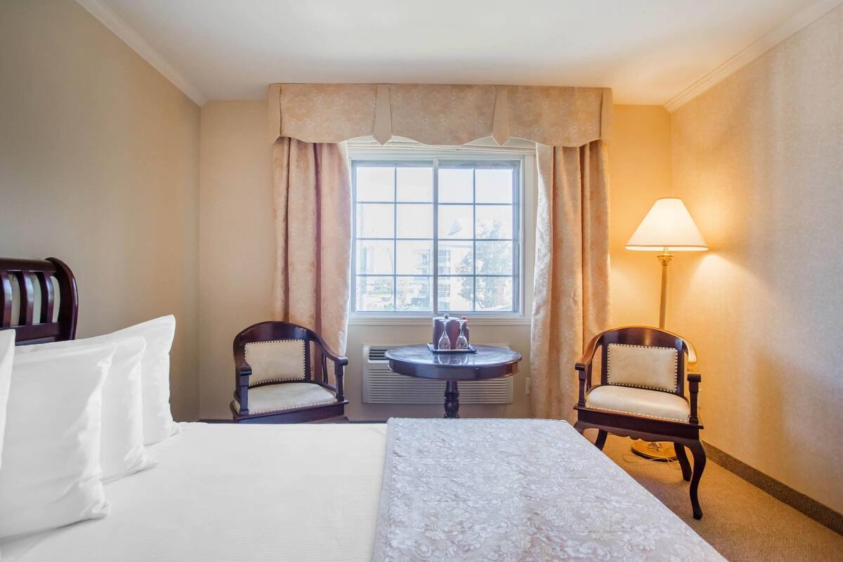 Una habitación de hotel con cama, dos sillones, una mesita con lámpara y ventana con cortinas.