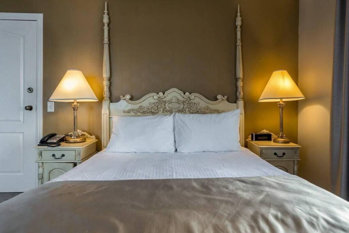 Una cama bien hecha con un cabecero clásico, dos mesitas de noche con lámparas y una puerta a la izquierda.