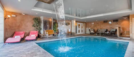 An indoor swimming pool with a waterfall feature, surrounded by red lounge chairs and wooden benches.