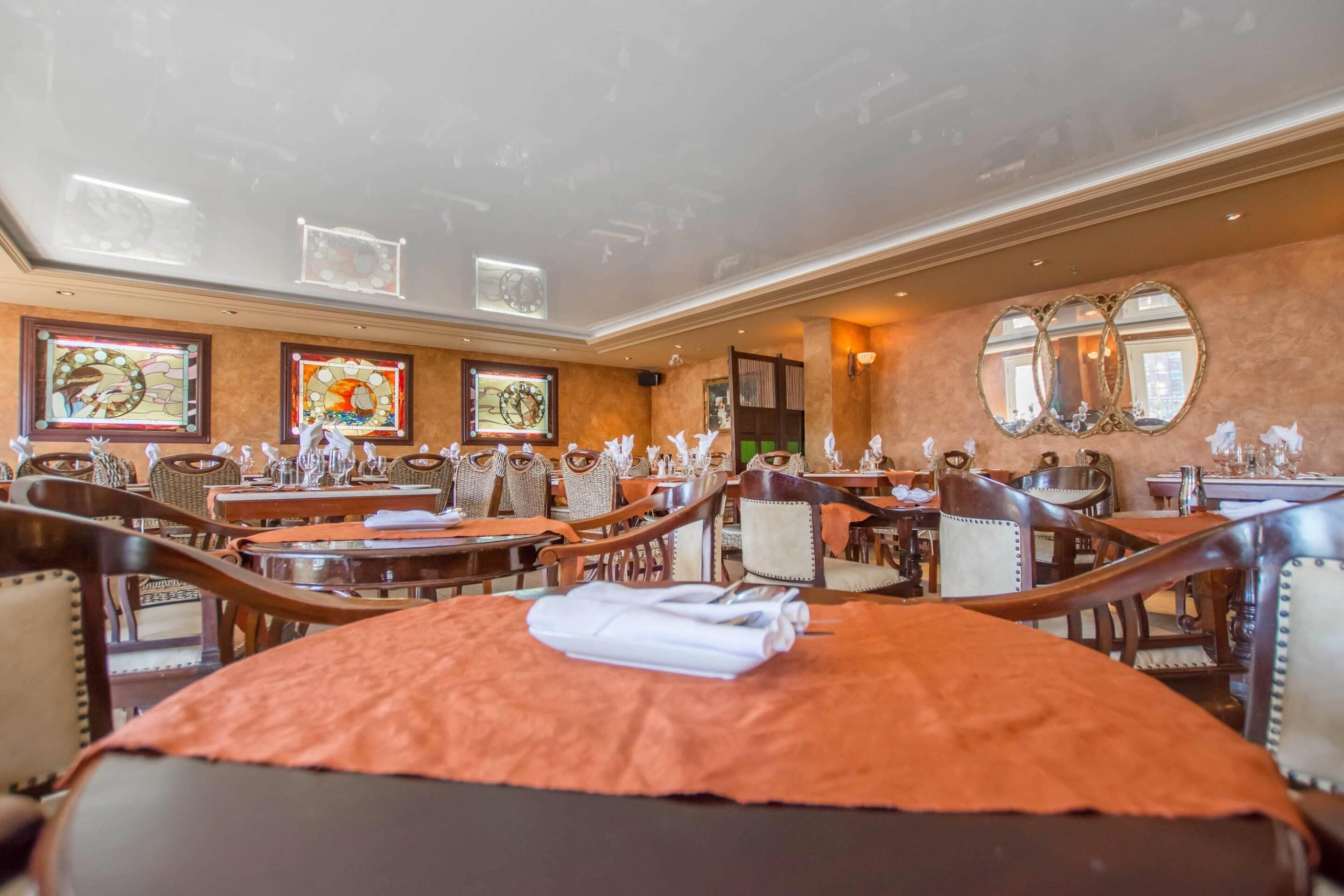 A hotel dining area with multiple tables and chairs, framed artwork on the walls, and a large mirror.
