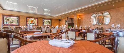A hotel dining area with multiple tables and chairs, framed artwork on the walls, and a large mirror.