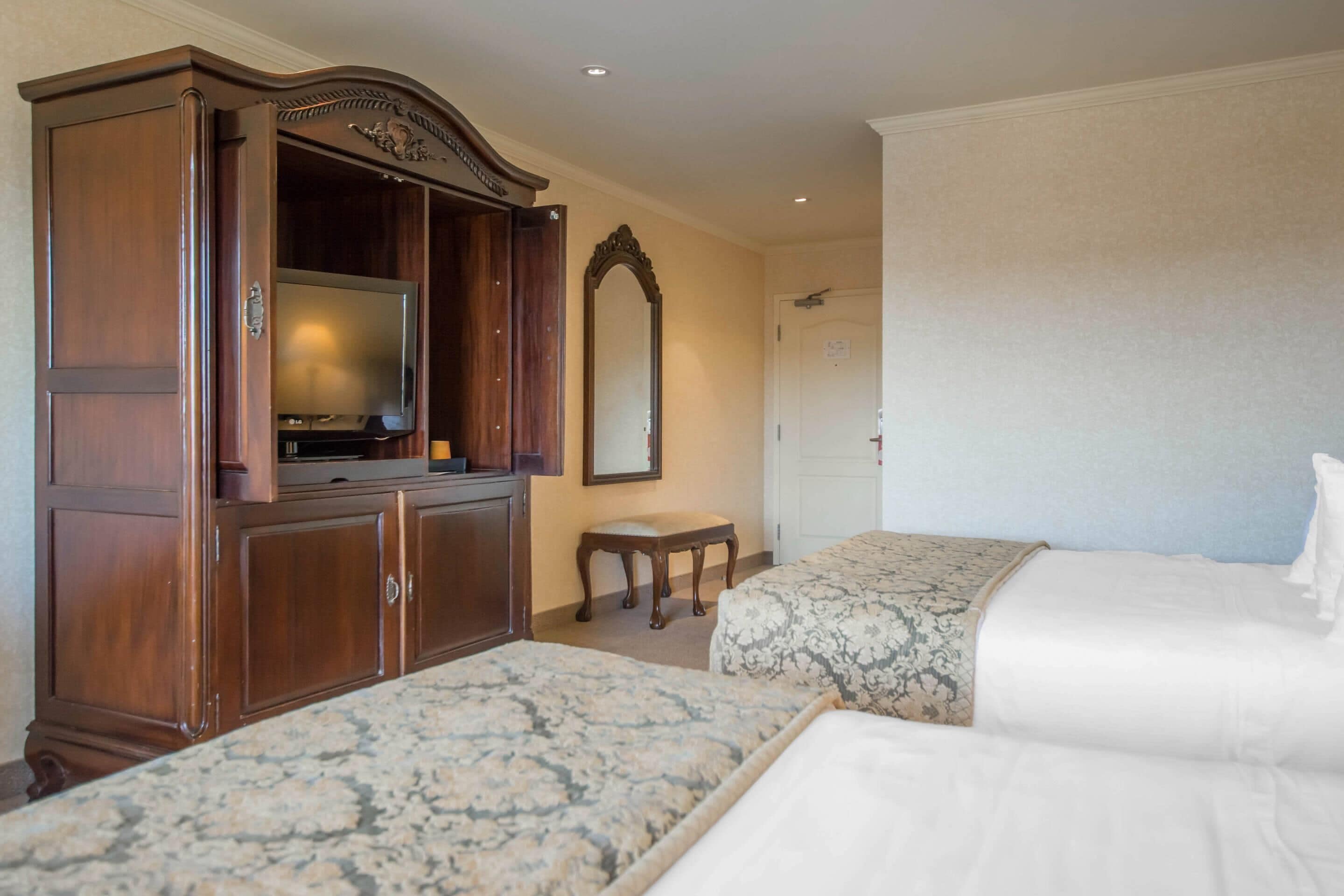 A hotel room with two beds, a wooden wardrobe, a television, a mirror, and a small table.