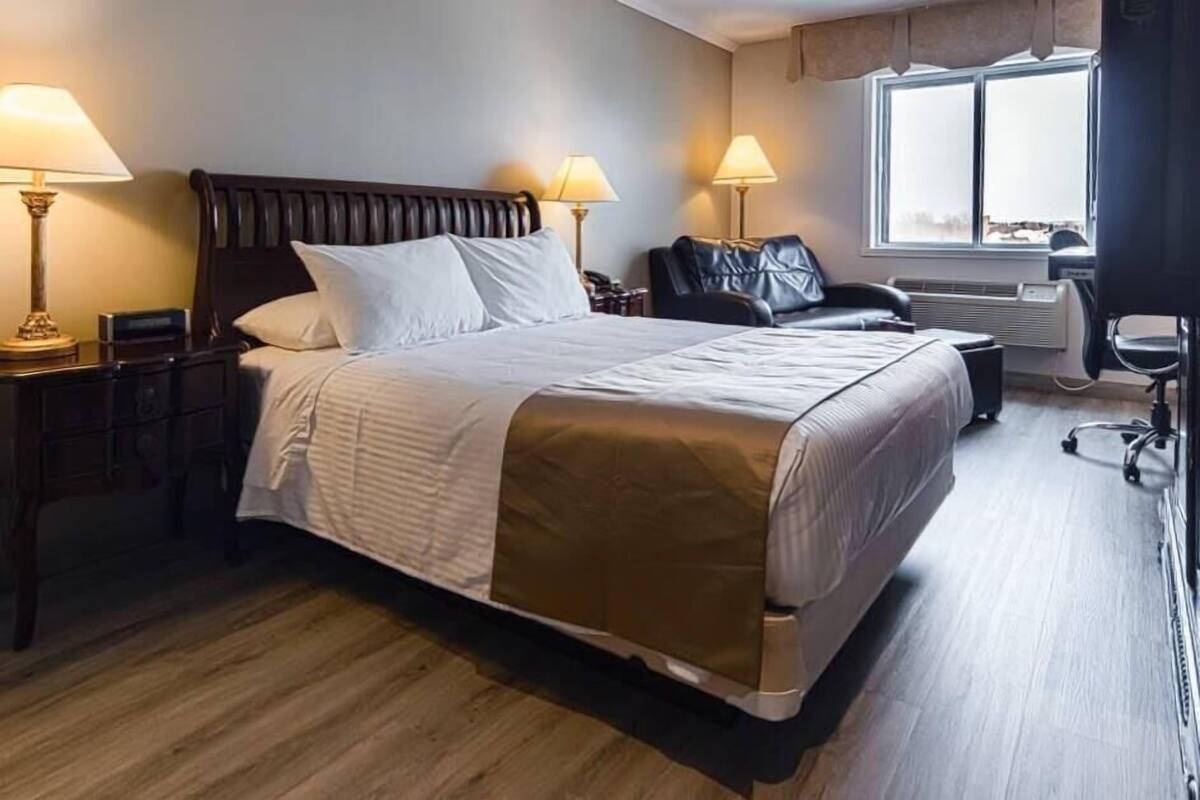 Habitación de hotel con una cama grande, dos mesitas de noche con lámparas, un sillón de cuero, un escritorio con silla y una ventana con vistas.