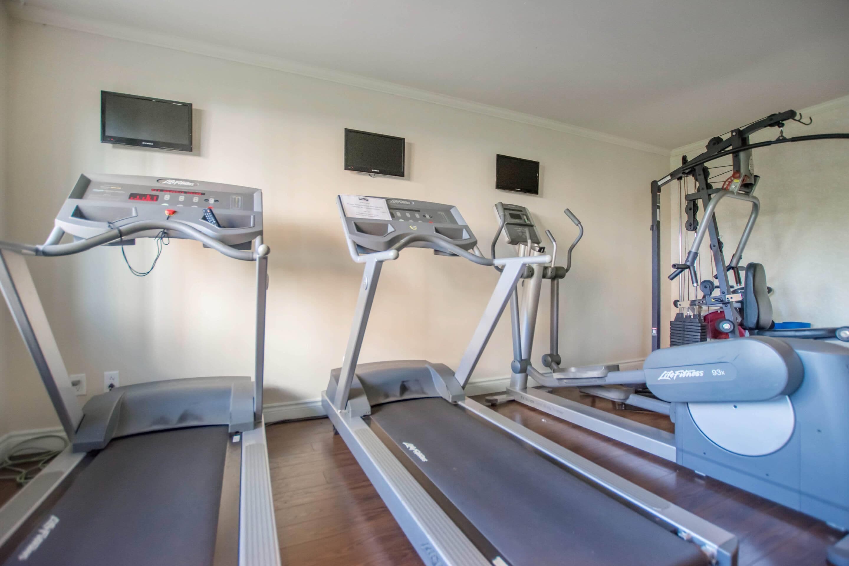 Tres cintas de correr en una sala con piso de madera, un televisor montado en la pared y una máquina de pesas.