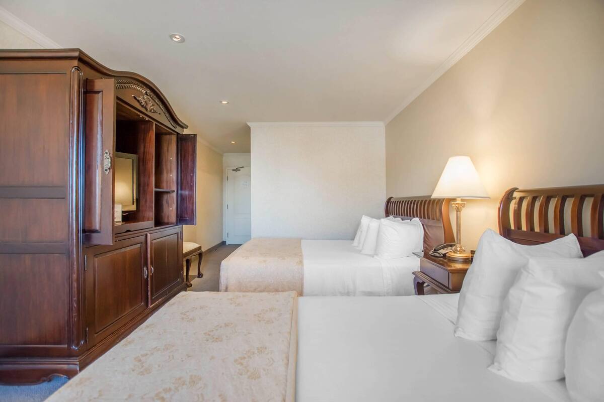 Habitación de hotel con dos camas, un armario de madera, una mesita de noche con lámpara y vista al pasillo.