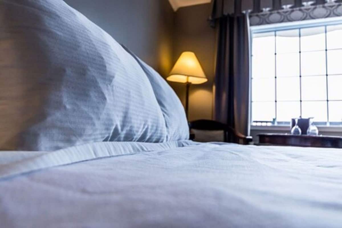 Una habitación de hotel con cama, lámpara, silla y ventana con cortinas.