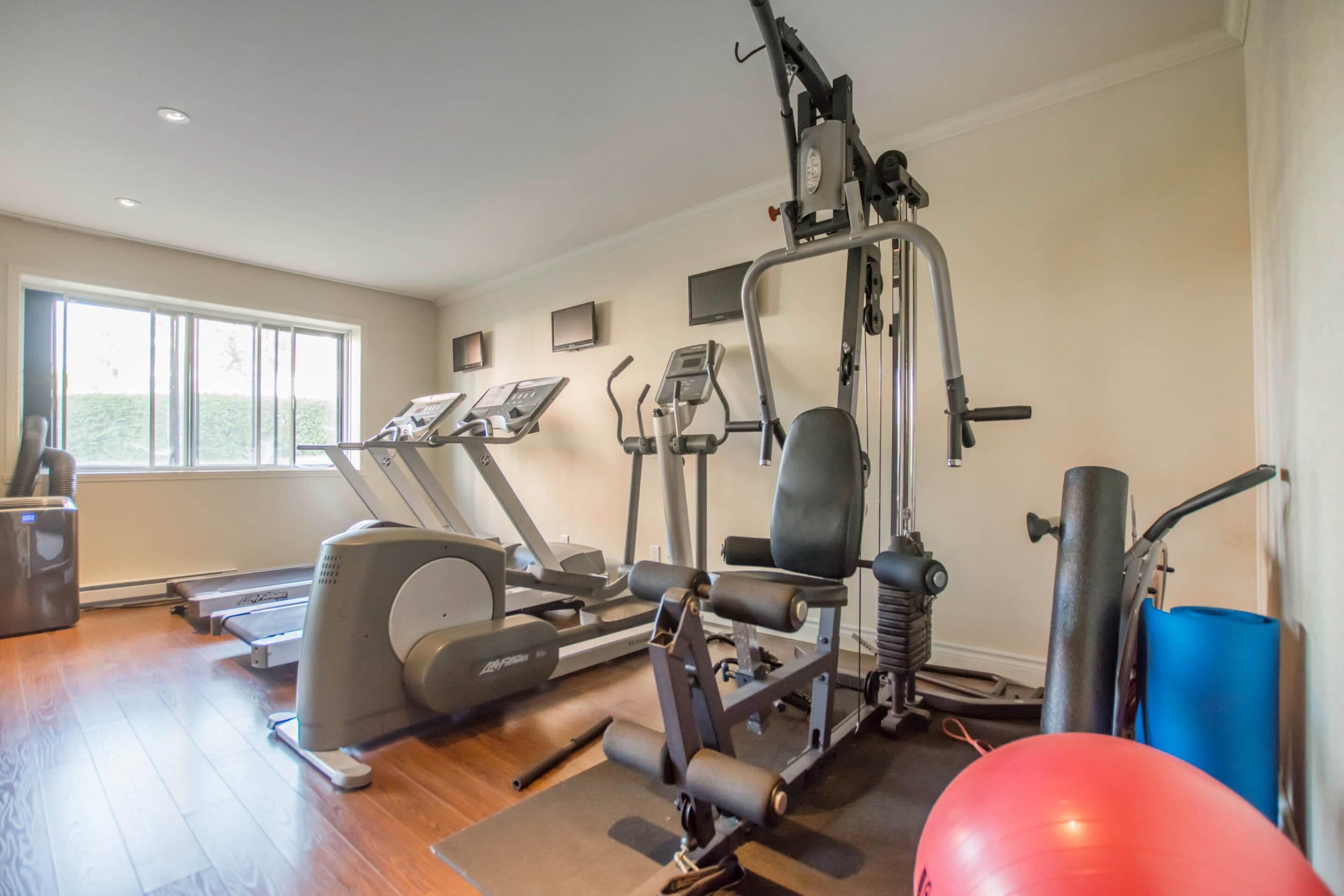 Un gimnasio en casa con máquinas de cardio, equipo de entrenamiento con pesas y una pelota de ejercicios roja.