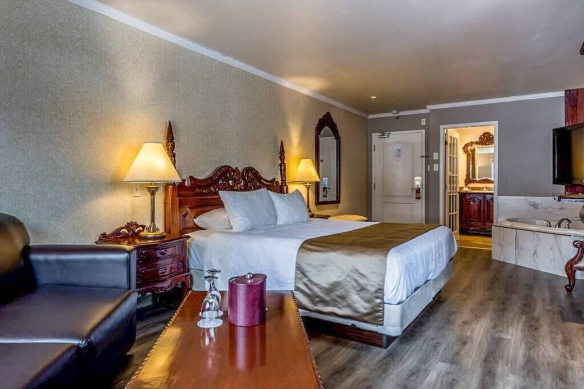 Habitación de hotel con una cama grande, dos lámparas de noche, una mesita de madera, una pequeña mesa con un vaso y un espejo sobre la cama.