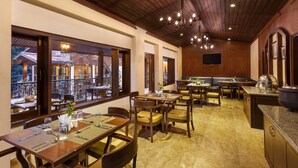 Un restaurant avec des tables et des chaises en bois, un espace cuisine et de grandes fenêtres donnant sur l’extérieur.
