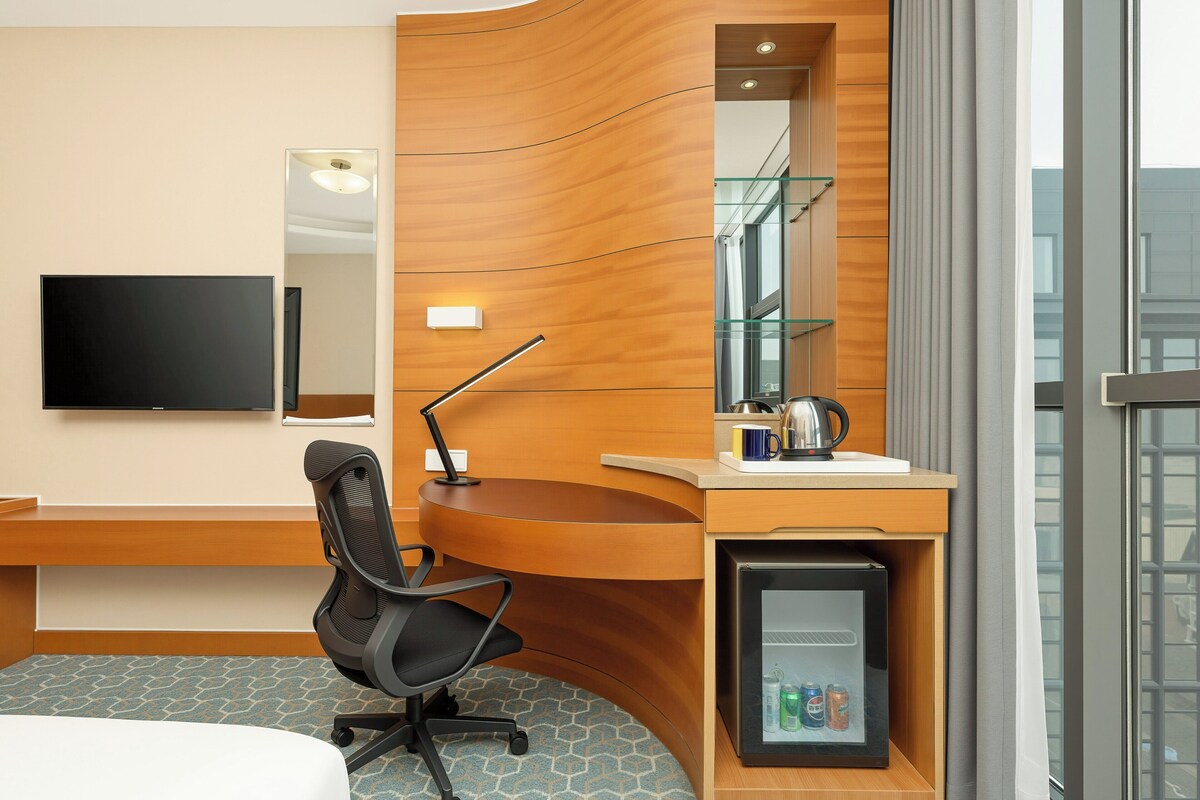 A hotel room with a curved wooden desk, a black office chair, a flat-screen TV, a minibar, and a large window.