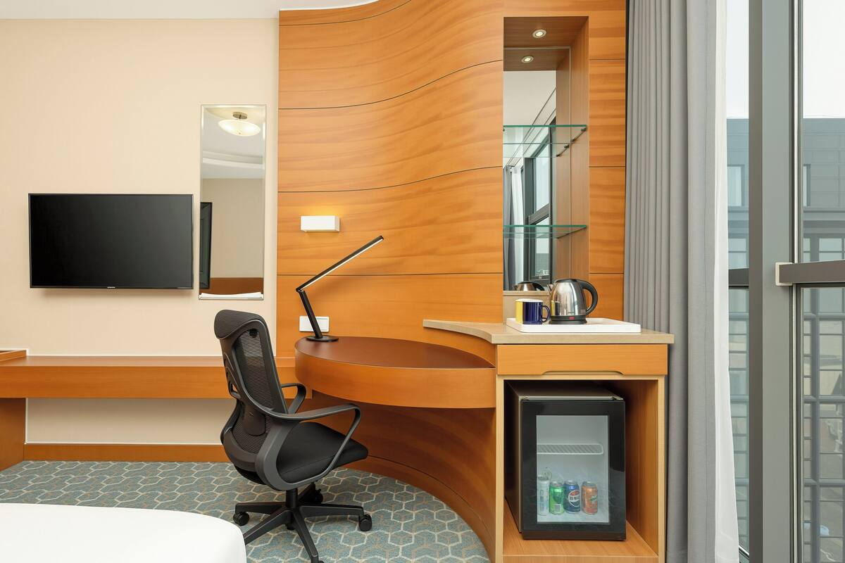 A hotel room with a curved wooden desk, a black office chair, a flat-screen TV, a minibar, and a large window.