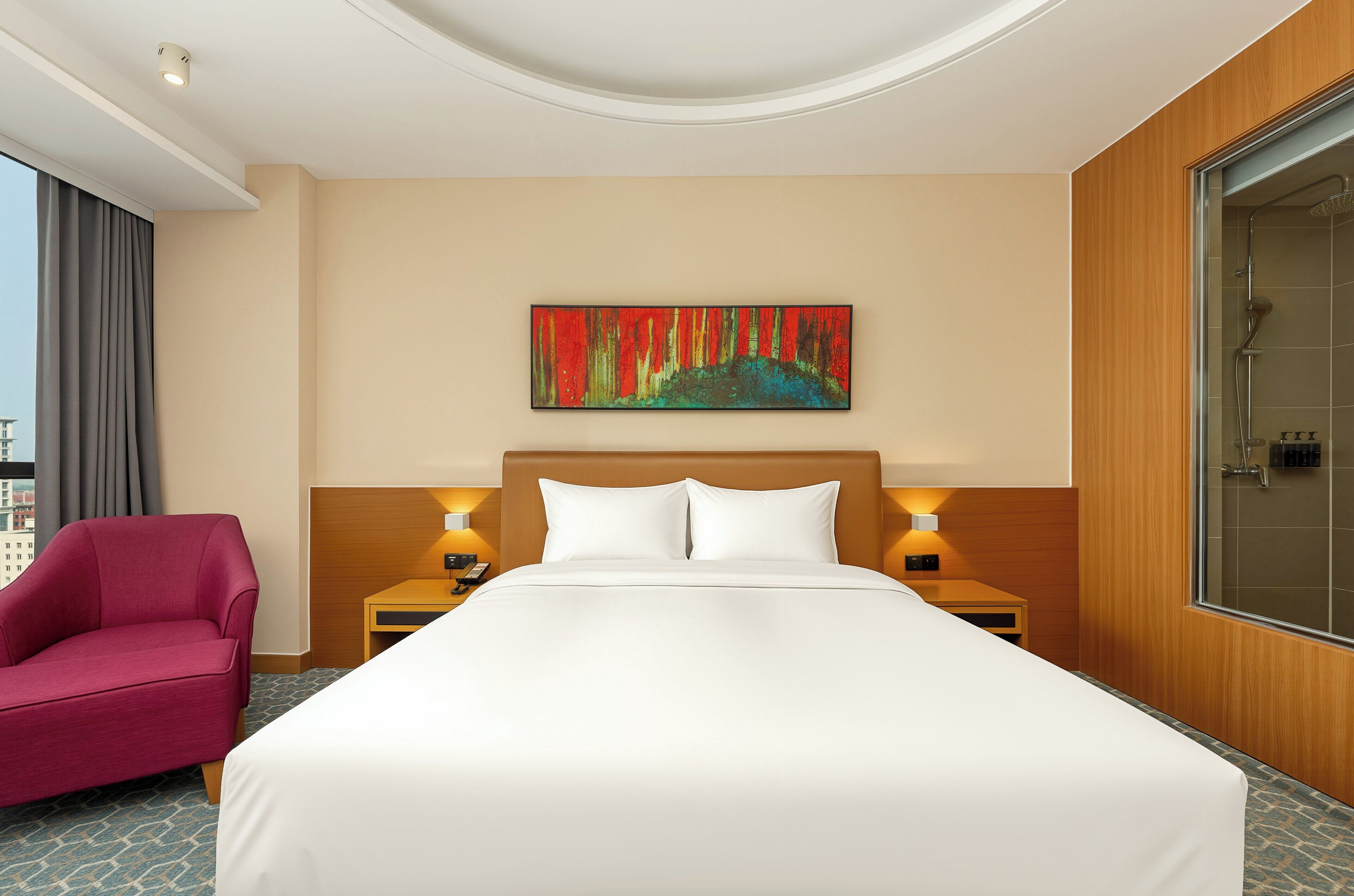 Een hotelkamer met een groot bed, een rode fauteuil, een schilderij aan de muur en uitzicht op de stad.
