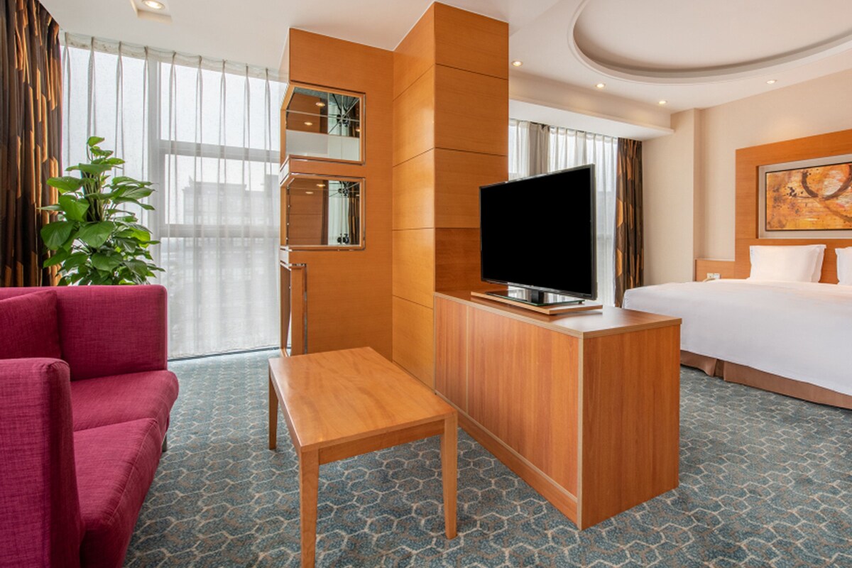 A modern hotel room with a large bed, a TV on a wooden stand, a desk, a chair, and a large window with curtains.