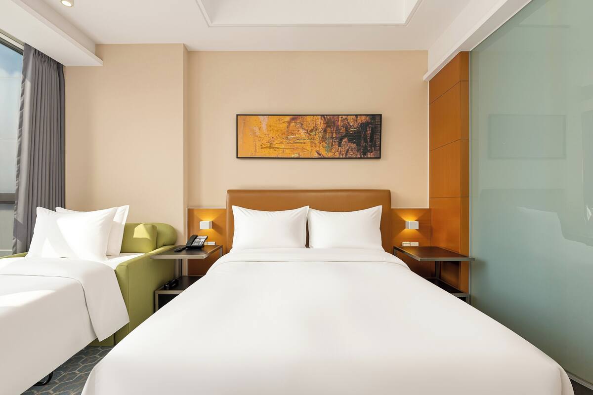 A hotel room with a large bed, two bedside tables, a green chair, a painting on the wall, and a window with curtains.