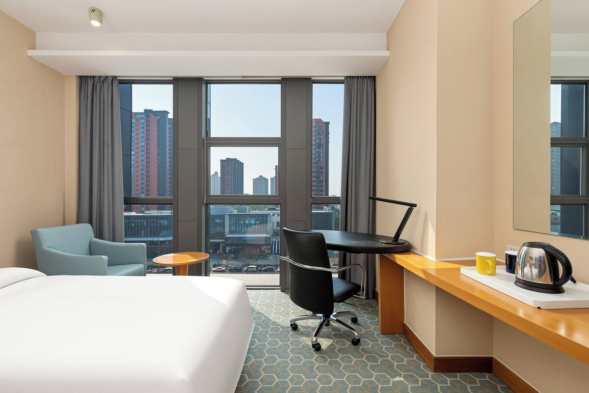 A hotel room with a bed, a desk, a chair, a small table, a kettle, and a view of tall buildings.