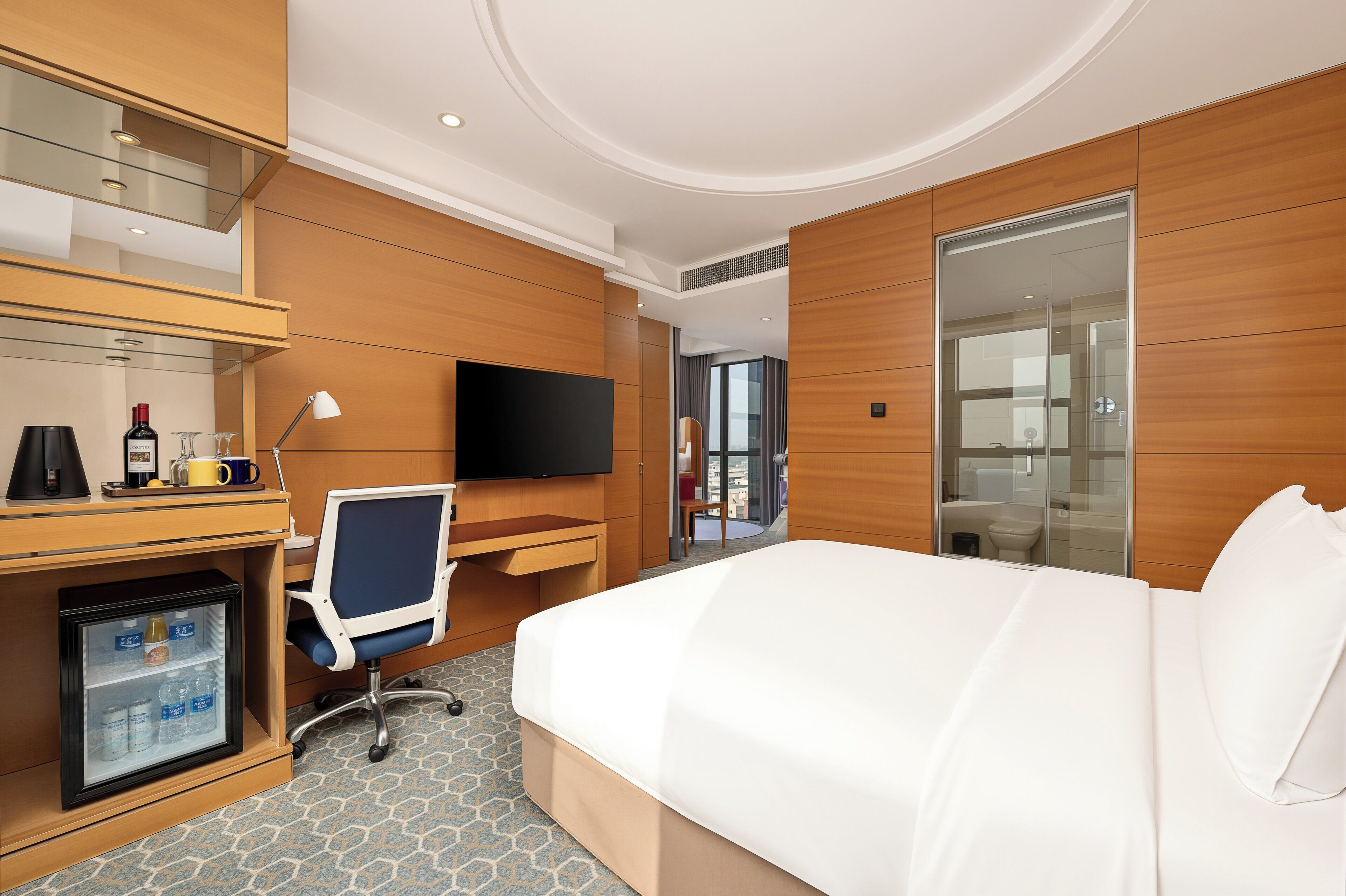 A hotel room with a bed, a desk with a chair, a television, and a minibar.