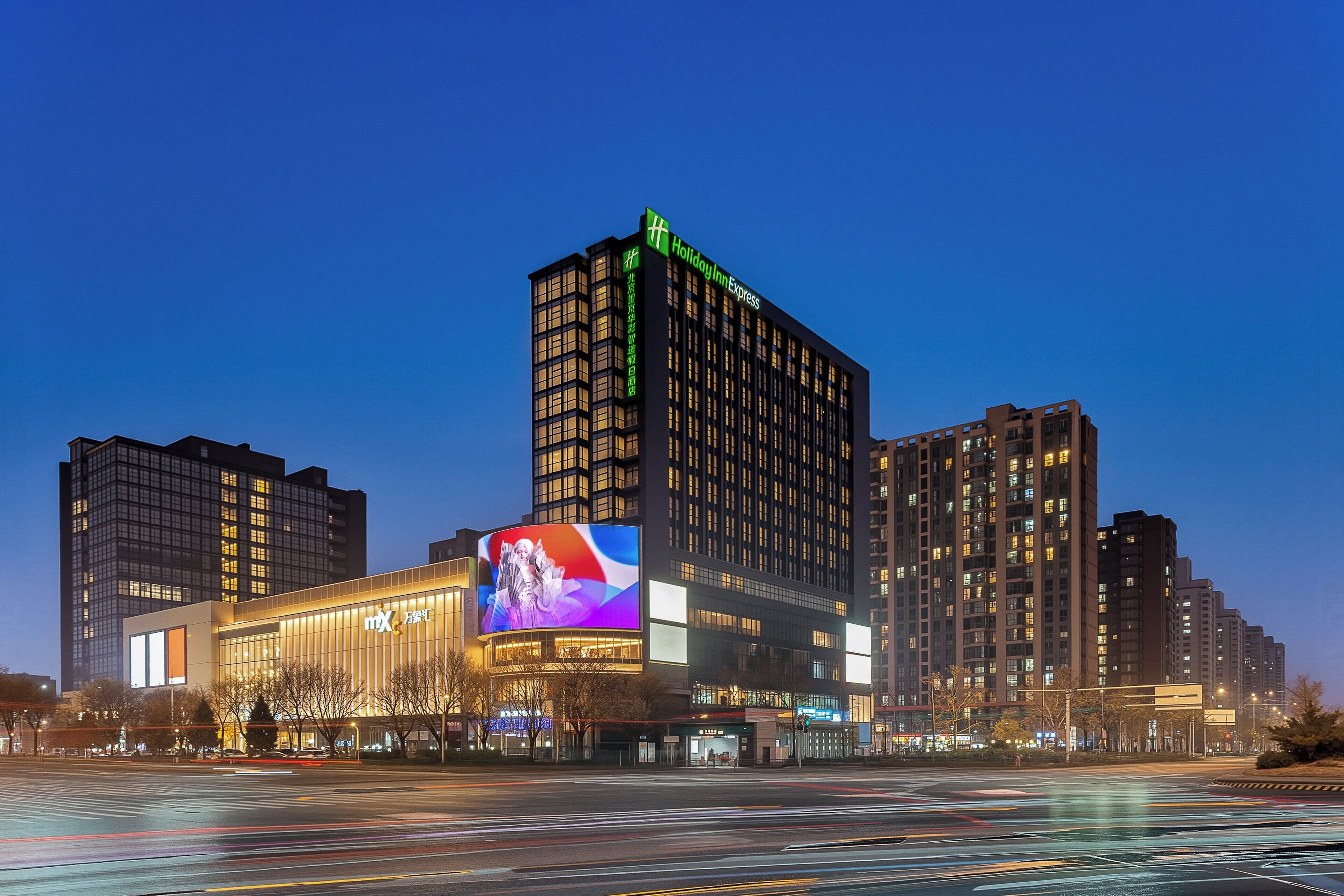 A modern hotel with a prominent digital billboard, high-rise buildings, and a well-lit urban street.