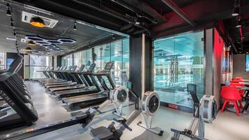 A gym with treadmills, rowing machines, and a view of a swimming pool through large windows.