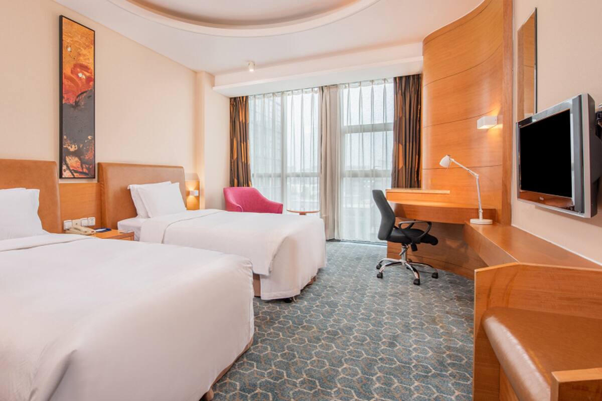 A hotel room with two beds, a desk, a TV, and a large window with curtains.