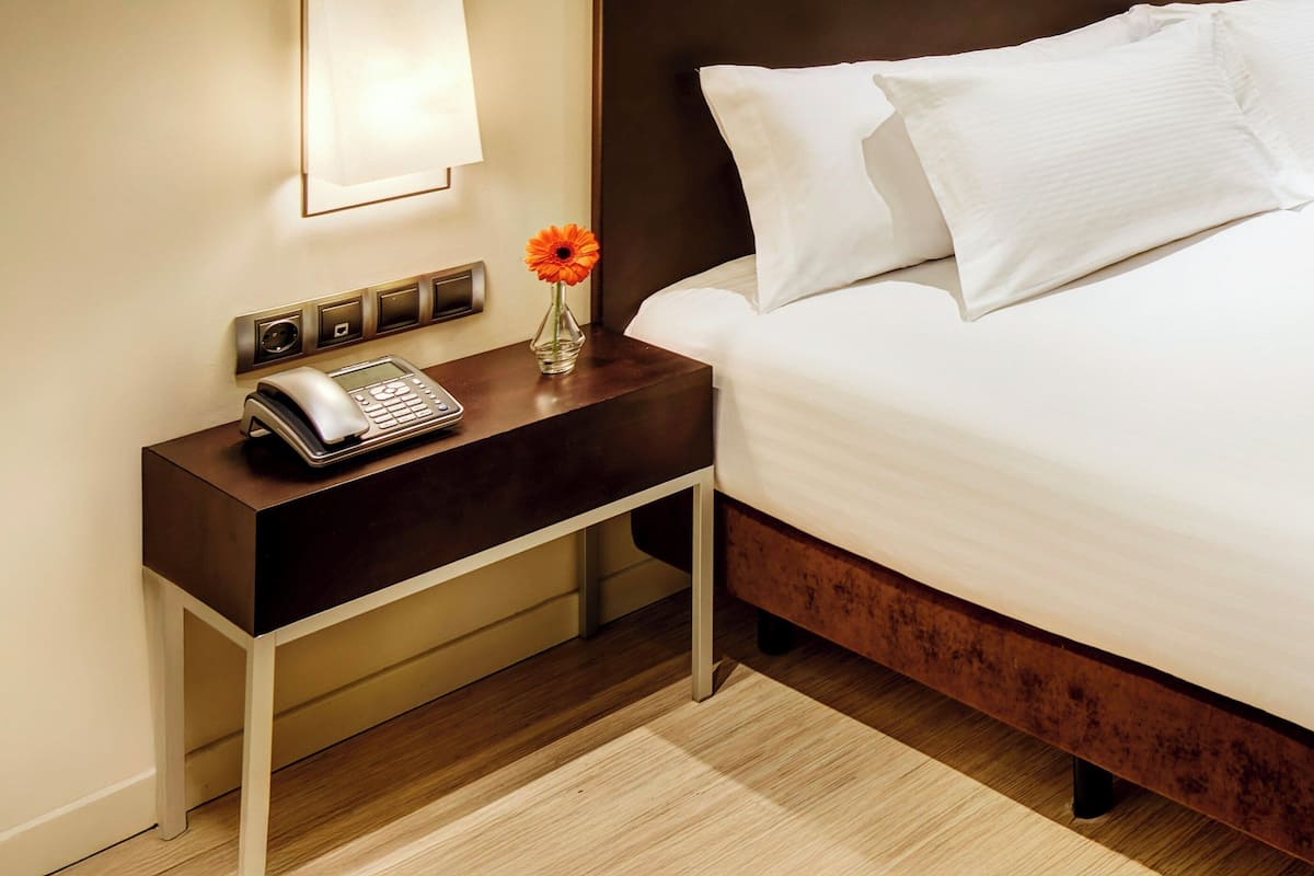 Habitació d'hotel amb una tauleta de nit de fusta, un telèfon, un vas amb una flor i un llit amb roba de llit blanca.