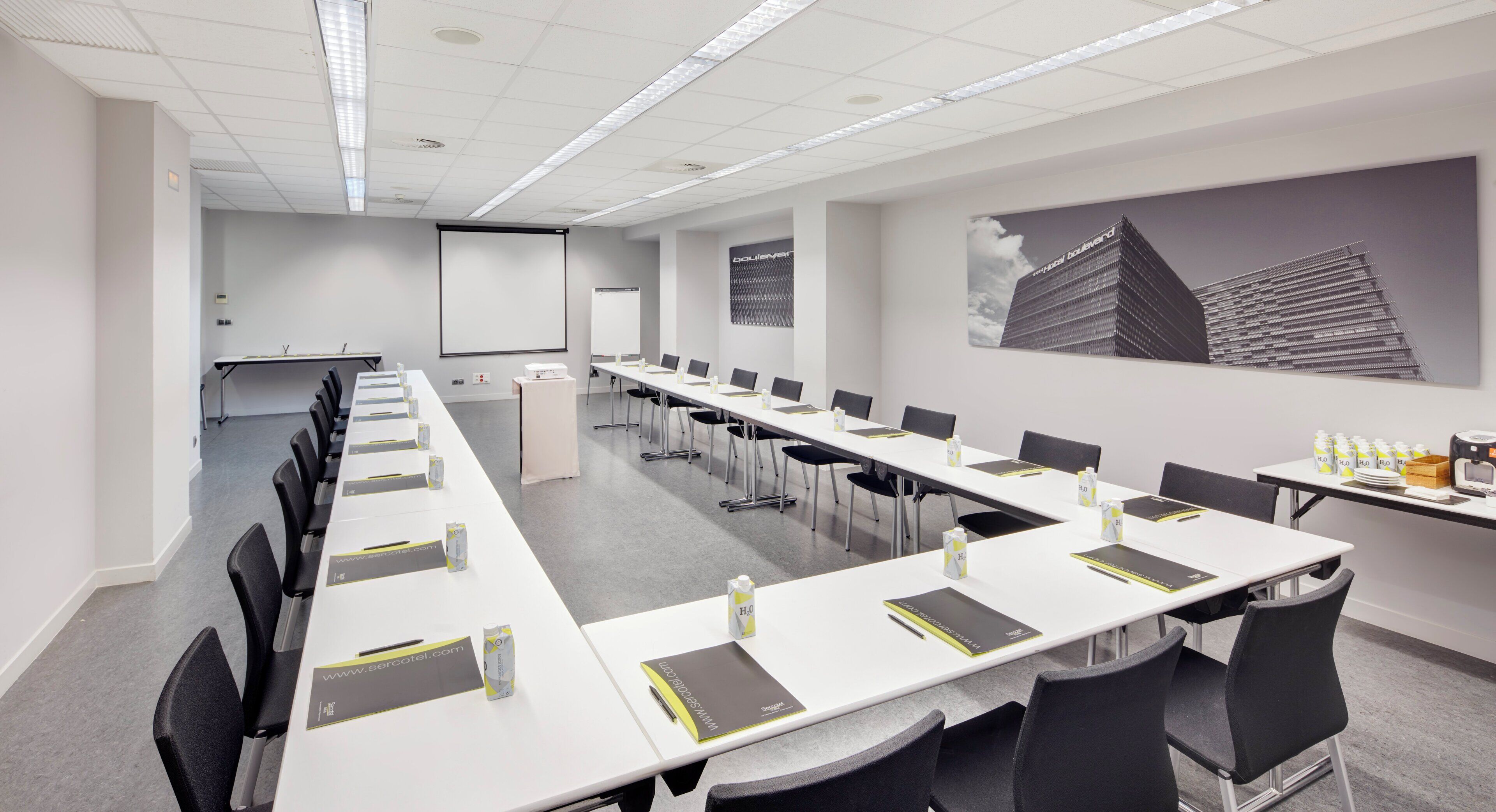 Een vergaderzaal met lange tafels, stoelen, een projectiescherm en een presentatiescherm.