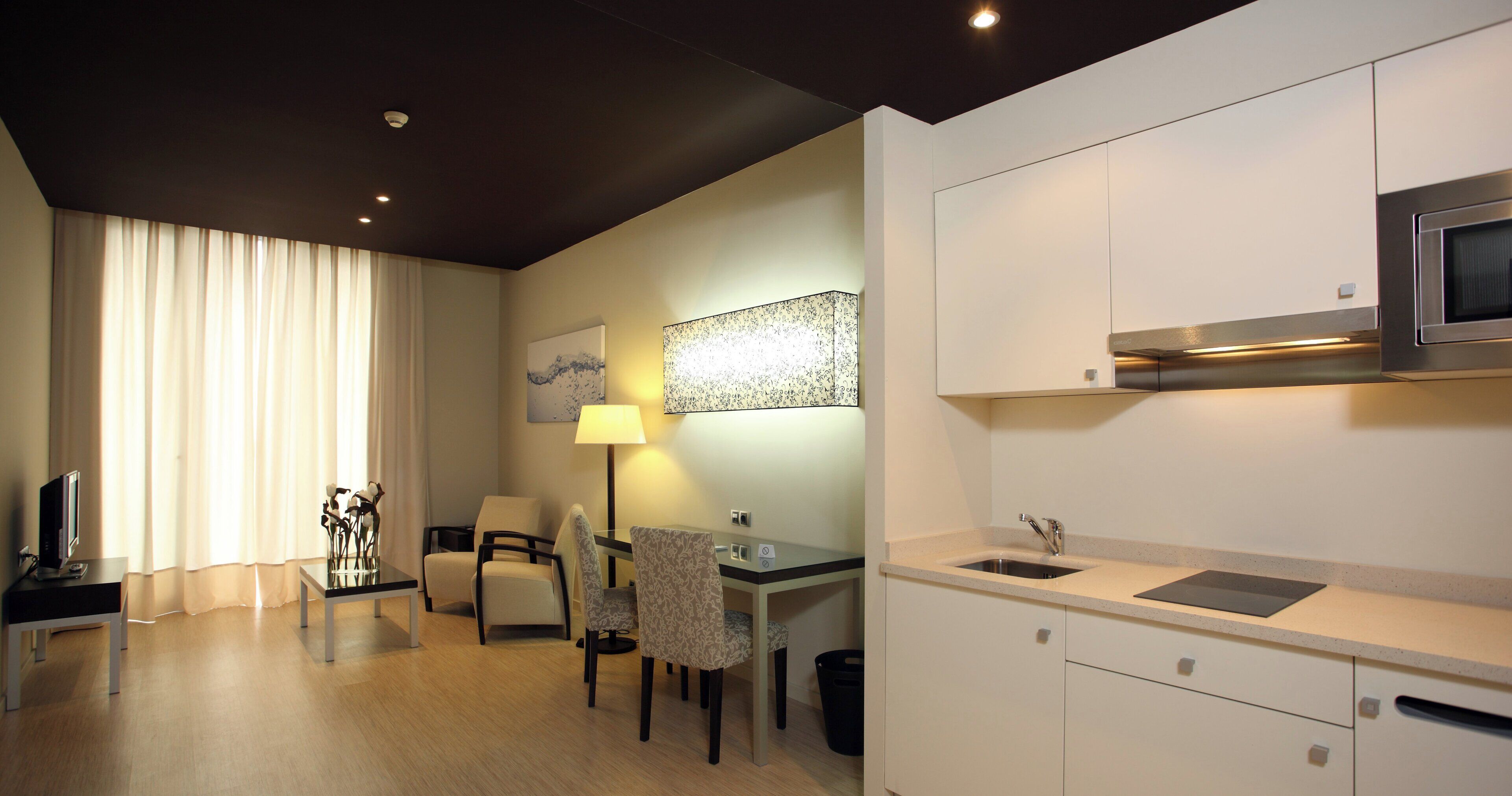 A modern hotel room with a dining area, a flat-screen TV, a lamp, and a large window with curtains.