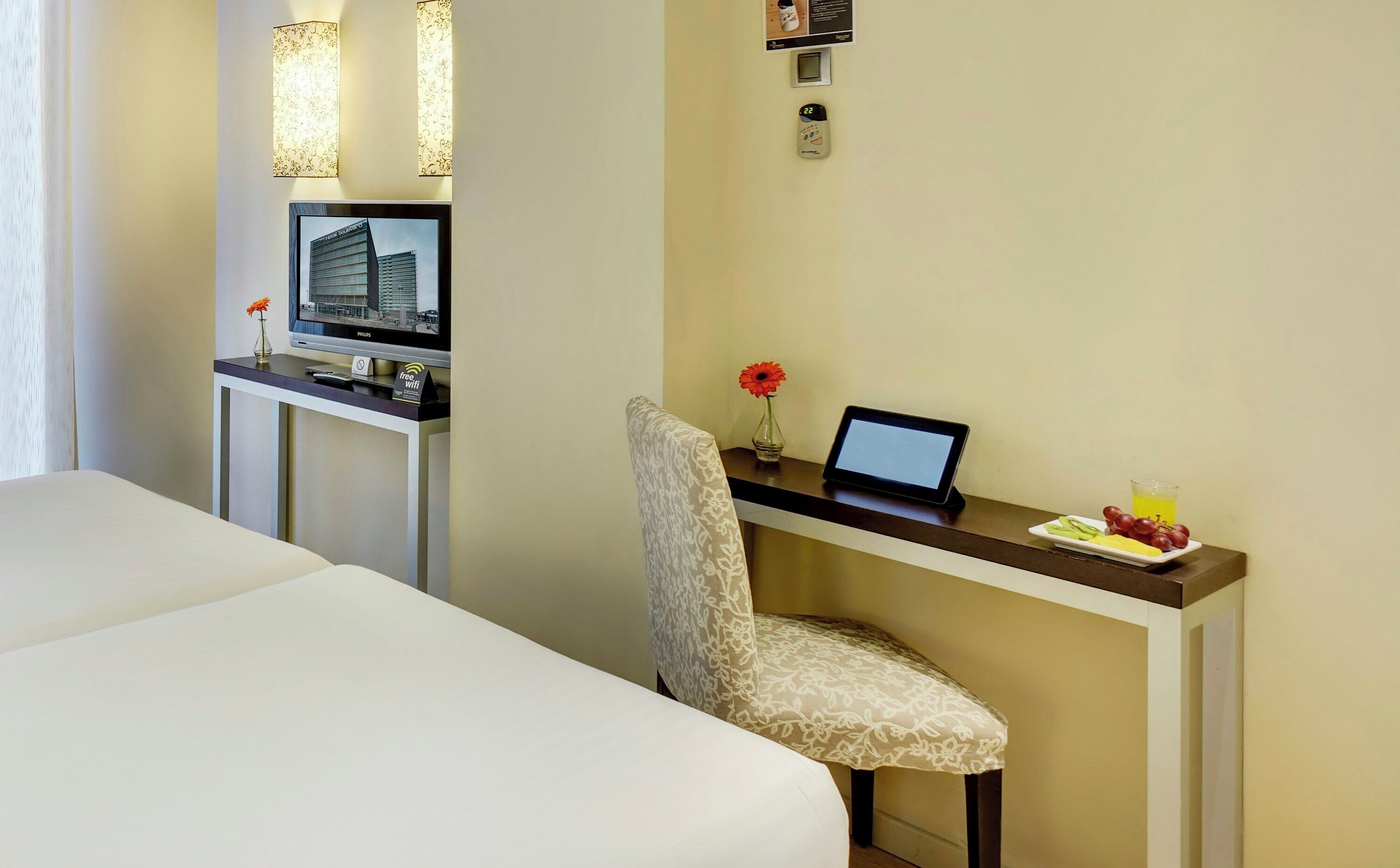 Habitación de hotel con cama, escritorio con una computadora portátil, florero con una flor y una bandeja de frutas.