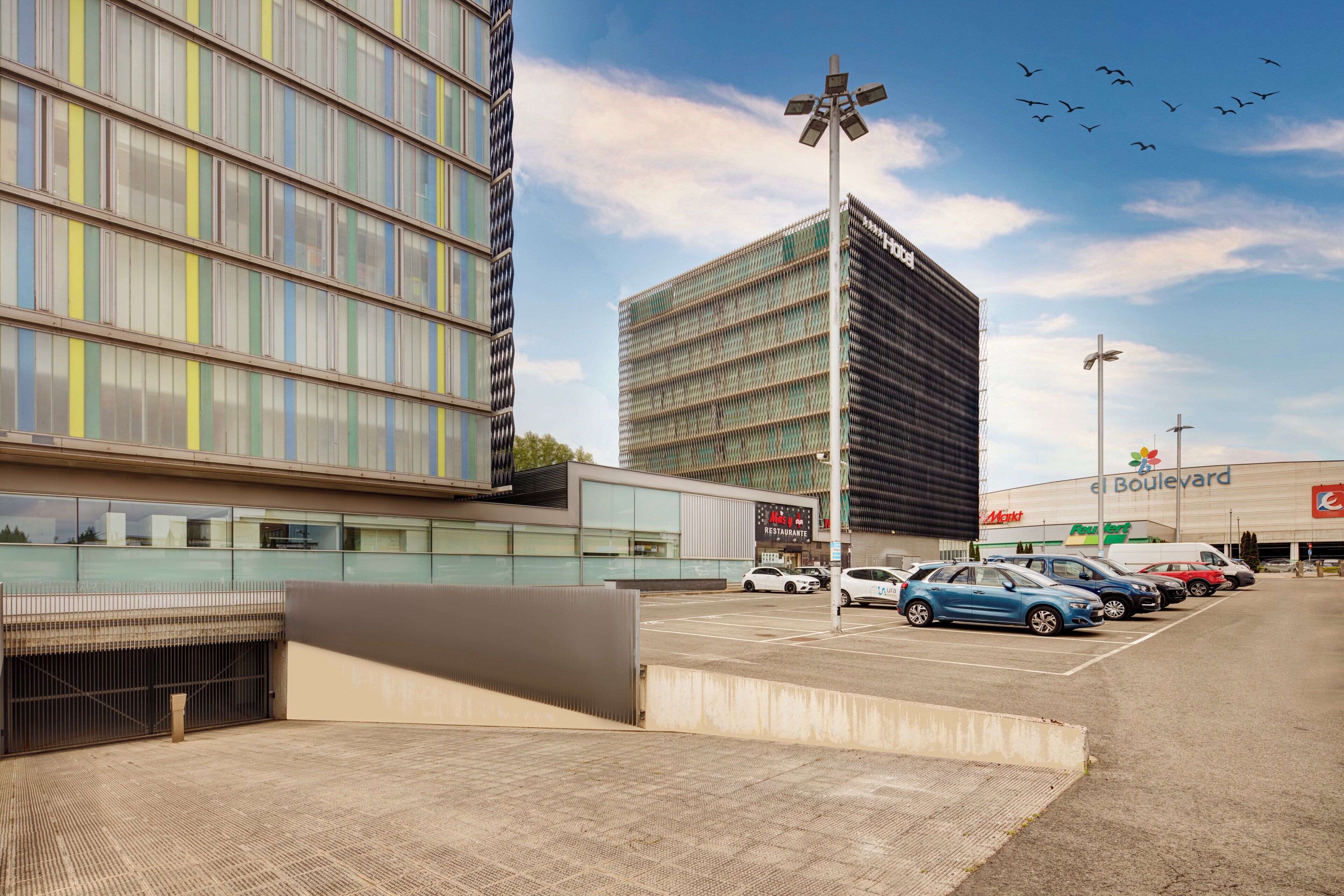 A modern building with a glass facade, a parking lot with several cars, and a clear sky with birds flying.