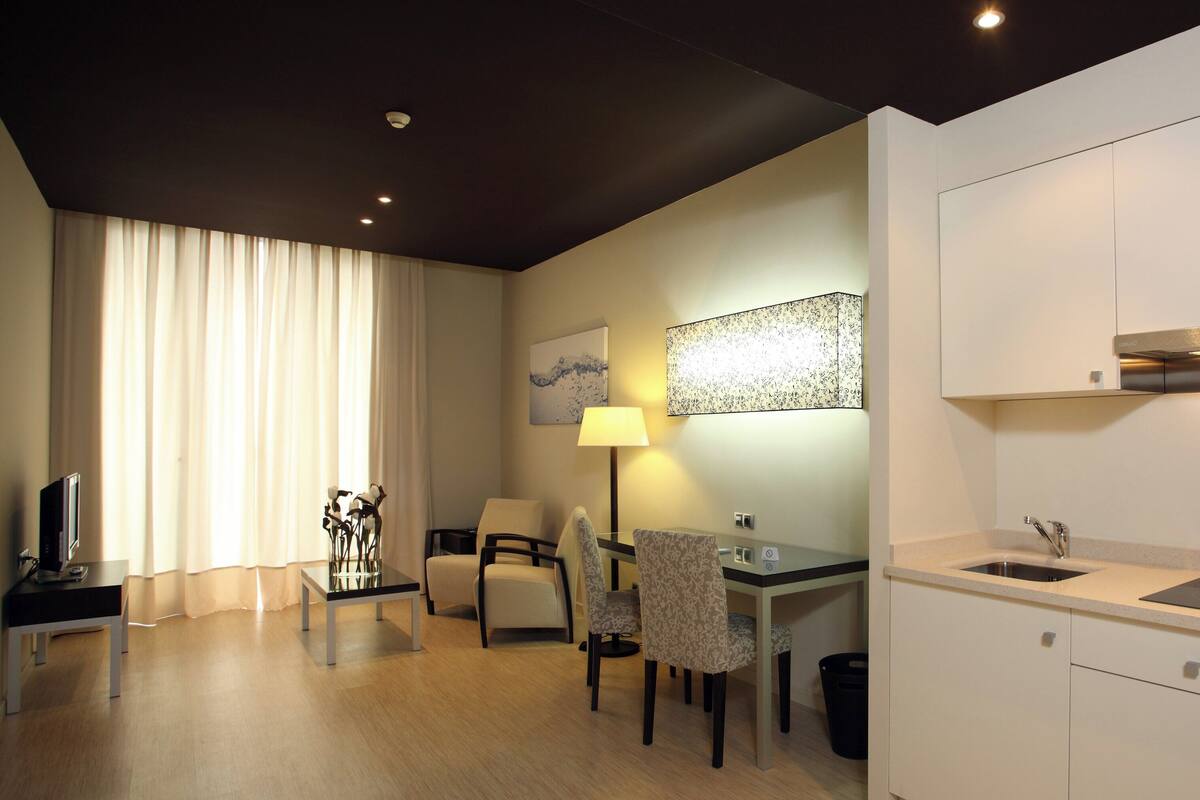 A modern hotel room with a dining area, a flat-screen TV, a lamp, and a large window with curtains.