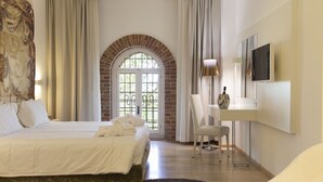A hotel room with a bed, a desk, a chair, a TV, and a large window with a brick arch.
