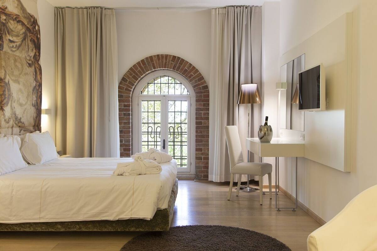 Une chambre d’hôtel comprenant un lit, un bureau, une chaise, une télévision et une grande fenêtre surmontée d’une arche en briques.