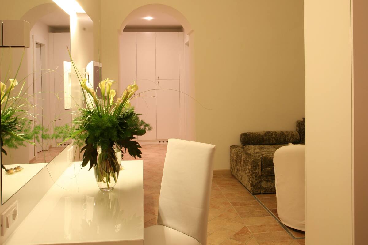 Un intérieur moderne avec un bureau blanc, une chaise et un grand miroir. On aperçoit une plante en pot sur le bureau et un canapé blanc à l’arrière-plan.