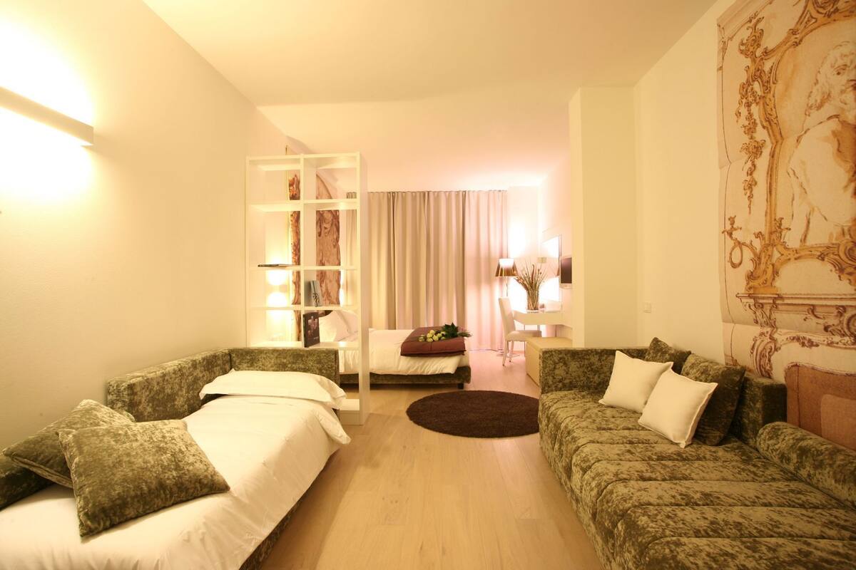Une chambre d’hôtel avec deux canapés verts à motifs, un lit avec une literie blanche, un plancher en bois, un bureau blanc avec une chaise, un petit tapis rond et un élément décoratif mural.