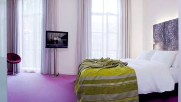 A hotel room with a bed, a television mounted on the wall, and a large window with sheer curtains.