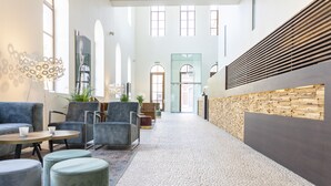 A spacious lobby with a high ceiling, seating area with sofas and chairs, a coffee table, and a decorative wall with a wooden panel. - Eburon Hotel - Different Hotels (Tongeren)